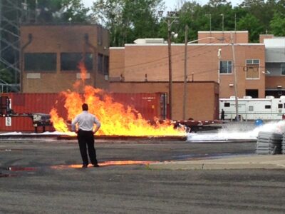 person stands in front of flames with building in background